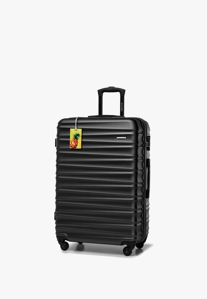 Valigia nera rigida con texture scanalata, manico telescopico, quattro ruote e un'etichetta per bagagli a forma di ananas colorato attaccata.