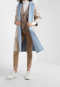 Abrigo azul claro de gran tamaño sobre un suéter beige, pantalones de pana marrones y zapatillas blancas. Notable contraste de colores y texturas superpuestas.