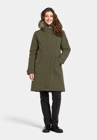 Oliven grøn, lang parka med pelsimitat på hætten, lynlås og klaplukning samt sidelommer. Bæres med sorte bukser og støvler.