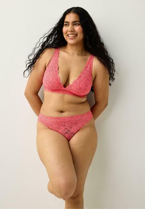 Kvinde med krøllet hår i pink blondetop og matchende undertøj står op ad en hvid væg, smilende med hænderne bag ryggen.