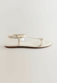 Sandale plate blanche avec fines lanières, boucle à la cheville et petits pendentifs ressemblant à des perles suspendus sur un fond uni.