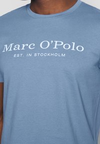 Camiseta de algodón azul con cuello redondo, que presenta el texto blanco "Marc O'Polo EST. EN ESTOCOLMO" en el frente. Textura suave, ajuste casual.