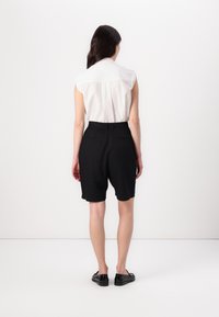 Schwarze, maßgeschneiderte Shorts mit lockerer Passform, kombiniert mit einem leichten weißen ärmellosen Shirt mit strukturiertem Muster. Schwarze Schuhe runden den Look ab.