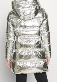 Metallisk silver puffer-jacka med huva, sydd mönster, elastiska partier och dragkedjestängning på sidan. Slät, glänsande yta.