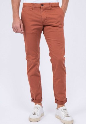 Homme portant un pantalon slim couleur rouille avec les ourlets retroussés et des baskets blanches, debout devant un fond uni.