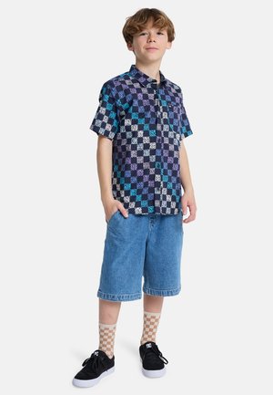 Chemise à manches courtes avec un motif à carreaux bleu marine, violet et bleu clair, associée à un short en denim et des baskets noires. Détail du tissu texturé.