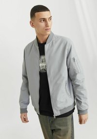 Graue Bomberjacke mit glatter Textur, gerippten Bündchen, seitlichen Reißverschlusstaschen und einem Reißverschluss. Über einem schwarzen Shirt mit Grafik getragen.