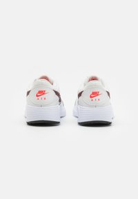 Bijele Nike tenisice s teksturiranim završetkom, s crvenim detaljima i crnom potplatom. "Nike Air" logo na peti. Zaobljen oblik.