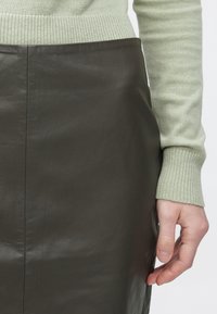 Temno zelena usnjena krilo z gladko teksturo, detajli na stranskem šivu, kombinirano s svetlo zelenim puloverjem z rebrastim manšetom, ki je viden na vrhu.