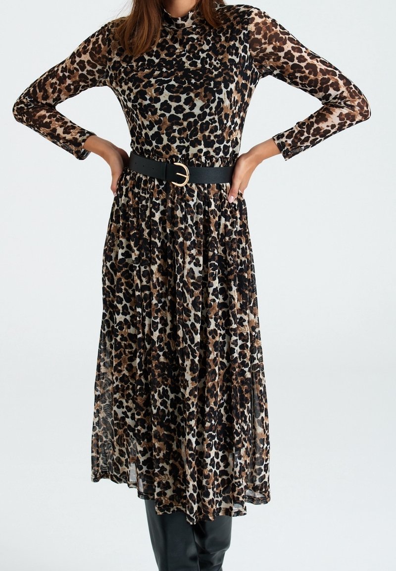 Femme portant une robe à manches longues et col haut à imprimé léopard, ceinture noire et bottes noires, debout les mains sur les hanches.