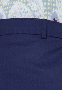 Gros plan sur la taille d’un pantalon bleu marine ajusté avec une passants de ceinture, porté avec un pull en maille à motifs bleu, jaune et blanc.