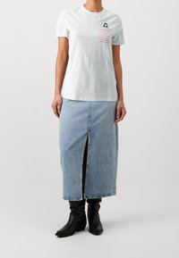 Kvinna som bär en vit t-shirt med en rödrandig ficka och en grafisk, denim midi-kjol med slits framtill, samt svarta cowboyboots.