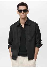Men's black jacket with a zip front, rolled sleeves, and a chest pocket, worn over a black knit shirt and light-colored pants.