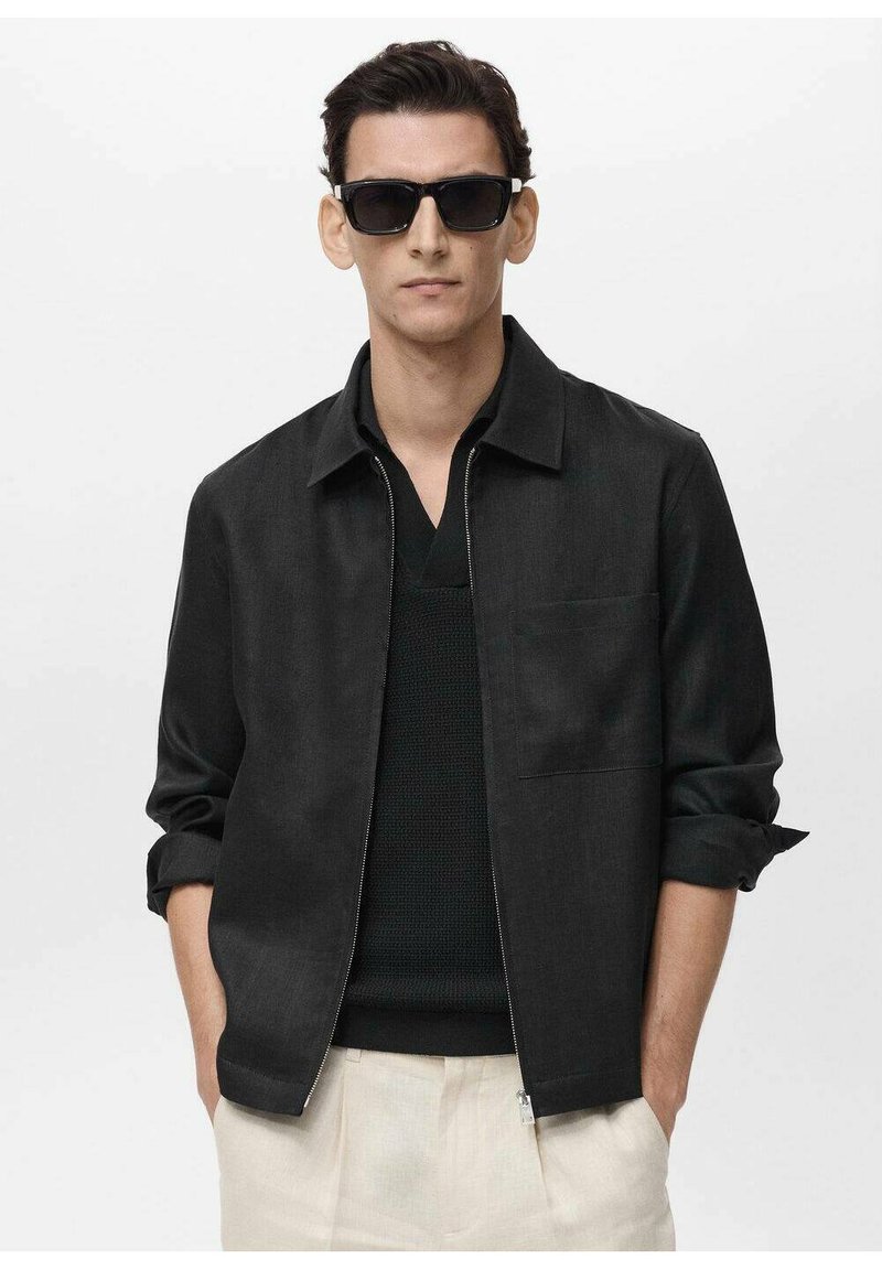Men's black jacket with a zip front, rolled sleeves, and a chest pocket, worn over a black knit shirt and light-colored pants.