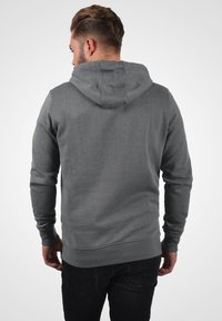 Haut à capuche gris avec une capuche à une seule couche, des poignets et un ourlet côtelés. Présente un petit logo sur l'épaule gauche. Matière en mélange de coton.