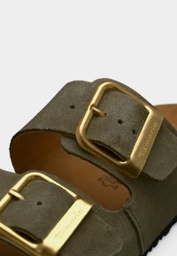 Sandale en daim vert avec deux larges brides, dotée de grandes boucles dorées et d'une semelle en liège brun, vue en gros plan des accents et de la texture.