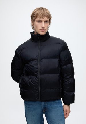 Homme blond portant une doudoune noire zippée, debout avec une main derrière le dos et un jean bleu devant un fond uni.
