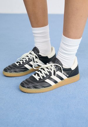 Personne portant des baskets Adidas Spezial noires et blanches avec des lacets blancs et des chaussettes blanches côtelées sur un sol bleu clair.