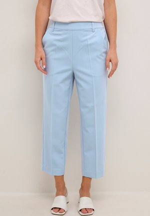 Pantaloni croppati blu chiaro con una vestibilità su misura, fronte piatto, tasche laterali e una texture liscia. Abbinati a mule bianchi con punta aperta.