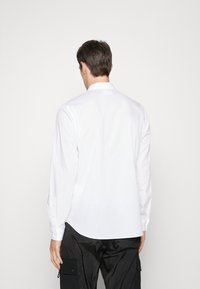 Camicia bianca a maniche lunghe con colletto button-down e una texture liscia, vestibilità su misura. Indossata con pantaloni neri dotati di tasche cargo.