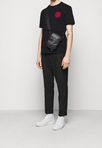 T-shirt noir avec un graphique rouge, pantalons noirs et baskets blanches. Un petit sac en bandoulière noir pend d'une sangle à travers la poitrine.