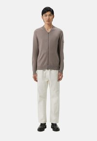 Ljusbrun zip-up kofta i strukturerad stickning, med V-halsringning, ribbade ärmslut och en sidoficka, ihop med vita byxor.