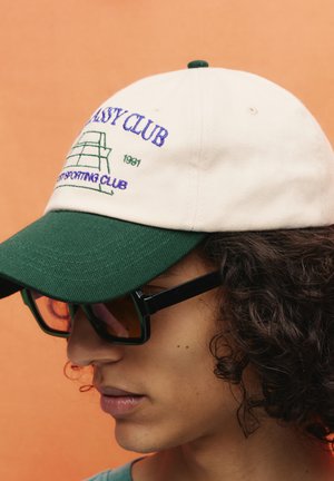 Beige Mütze mit grünem Schirm und verstellbarem Riemen, ausgestattet mit gesticktem Text und Grafiken in Blau und Grün. Getragen mit einer dunklen Sonnenbrille.