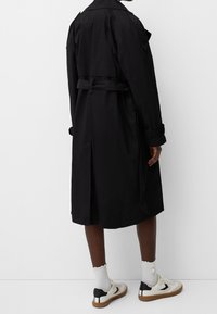 Czarny trenchcoat z paskiem w talii, długimi rękawami i rozcięciem z tyłu. Noszony na białe skarpetki i czarno-białe sneakersy. Gładka struktura.