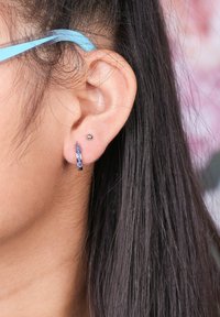 Zilverkleurige creool met een gestructureerd oppervlak en blauwe accenten, naast een kleine stud, gedragen aan het oor van een persoon.