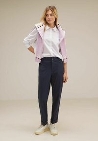 Gilet lavorato a maglia di un viola chiaro con motivi bianchi e bordeaux, indossato sopra una camicia bianca abbottonata e pantaloni navy su misura; sneakers beige.