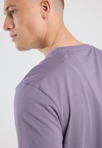 Junger Mann mit heller Haut, der ein lila Rundhals-T-Shirt trägt, von einem seitlich-hinteren Winkel vor einem schlichten hellen Hintergrund gezeigt.