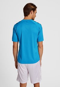 Koszulka sportowa z krótkim rękawem w jaskrawym niebieskim kolorze, z okrągłym dekoltem i czarnymi akcentami na ramionach, noszona z białymi szortami.