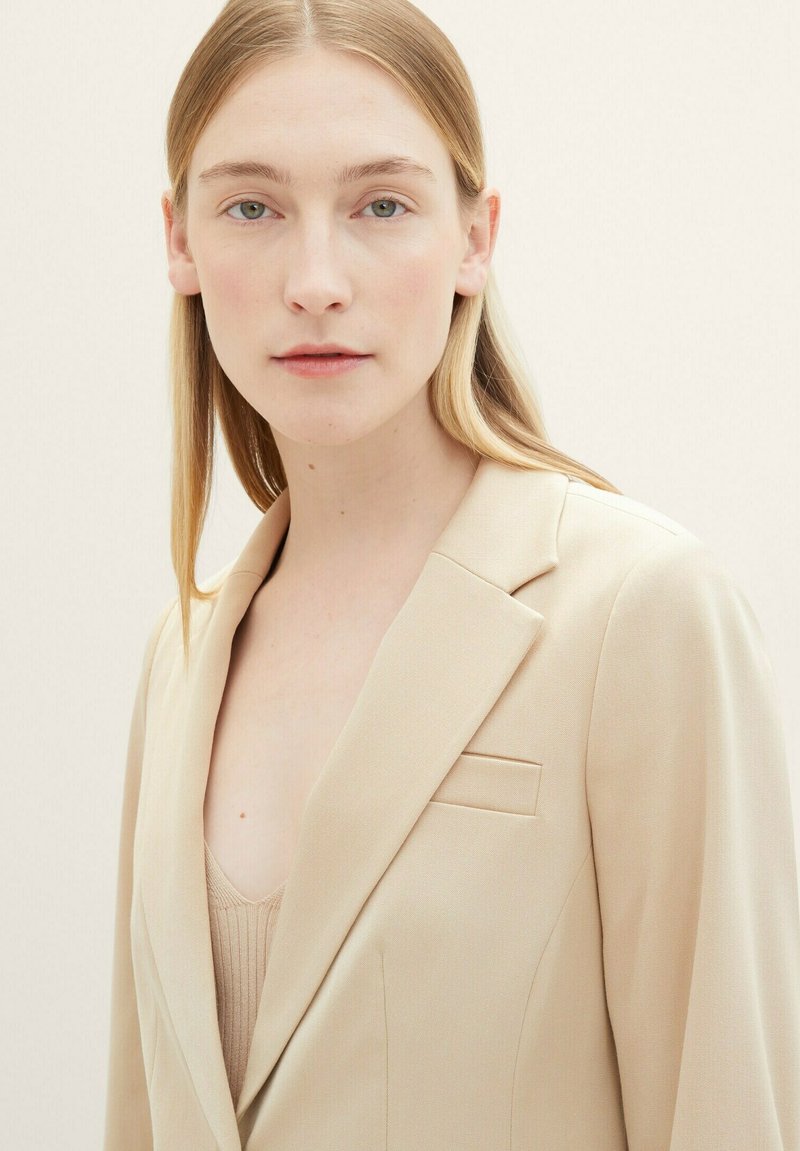 Blazer beige con rever a scannafisso, tasca sul petto singola e vestibilità sartoriale. Sotto c'è un top rigato beige. Sfondo neutro.