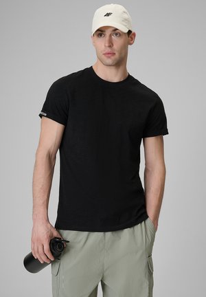 Mężczyzna w białej czapce i czarnym t-shircie trzyma czarną butelkę na wodę, ubrany w jasnoszare spodnie cargo, stoi na tle jednolitego szarego tła.