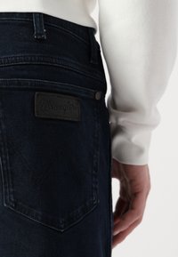Tamne traperice u tamnoj boji s kožnom oznakom Wrangler, koja se ističe kontrastnim šavovima, prilagođenim dizajnom i vidljivim oblikom džepa.