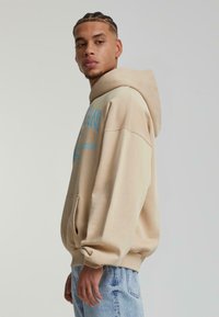 Felpa con cappuccio beige, maniche lunghe e tasca frontale. Presenta una scritta blu stampata sul petto. Indossata con jeans azzurri chiari.