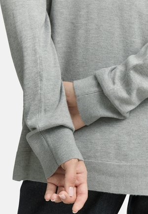 Manos con uñas naturales descansando detrás de la espalda, vistiendo un suéter de manga larga gris claro y pantalones oscuros, contra un fondo liso.