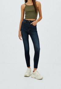 Jeans de mezclilla oscura de tiro alto, ajuste skinny con costuras naranjas y diseño clásico de cinco bolsillos, combinados con una camiseta sin mangas verde oliva y zapatillas blancas.