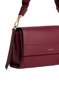 Borsa a mano in pelle granata con chiusura a patta, chiusura in metallo dorato, tracolla staccabile e piccolo logo dorato "Coccinelle" sul davanti.