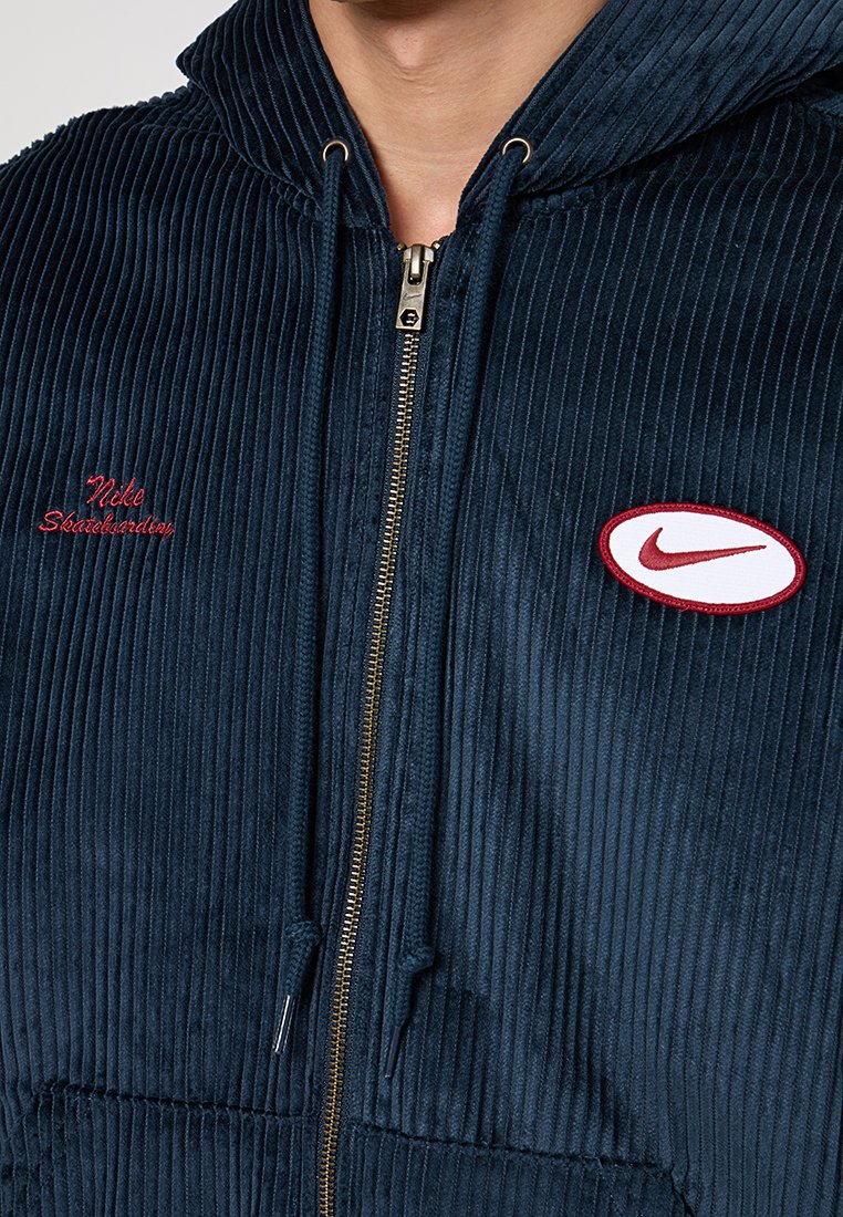 Haut de sweatshirt zippé en velours côtelé bleu marin avec une fermeture éclair dorée, brodé "Nike" en rouge, et un écusson blanc avec un contour rouge. Tissu texturé.