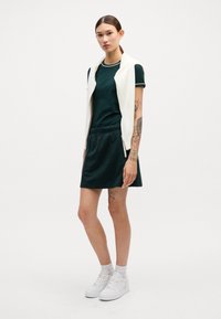 Vestido verde de manga corta con cuello blanco, combinado con un suéter sin mangas blanco y zapatillas blancas. El modelo tiene tatuajes visibles en los brazos.