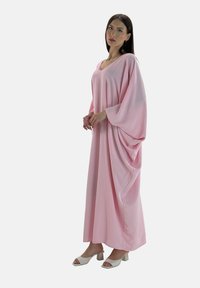 Lange roze jurk met een V-hals, losse mouwen en geplooide details aan de zijkanten. Gepaarde met witte block-heeled sandalen. Zachte textuur.