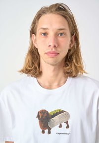 White cotton t-shirt featuring a graphic of a brown dog carrying a gray sandwich, with green accents. Logo text below the design.