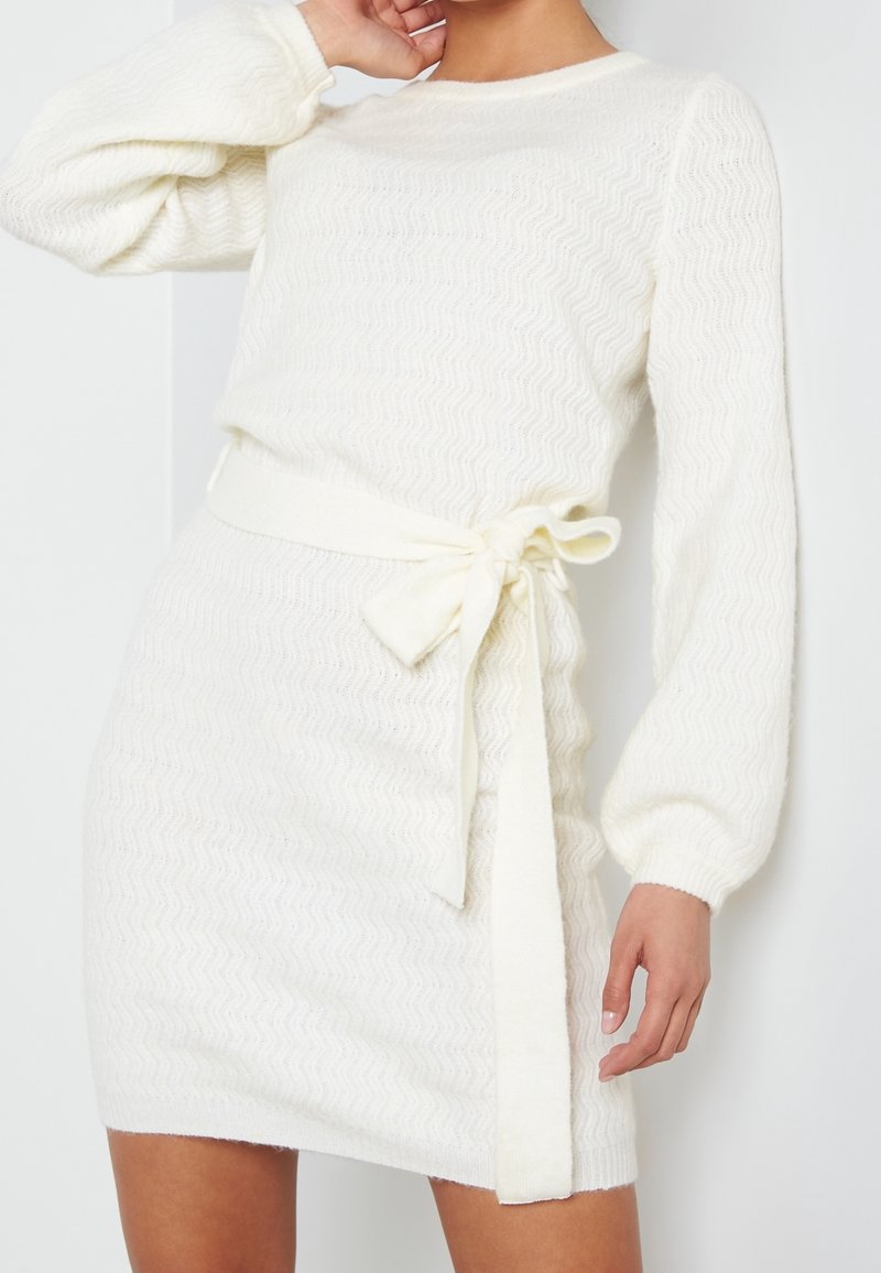 Robe en tricot blanc avec un motif en zigzag texturé, manches longues bouffantes et ceinture nouée à la taille. Matière douce avec un léger éclat.
