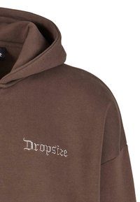 Smeđa dukserica napravljena od mekog materijala, sa malim izvezenim logotipom u dekorativnom fontu na prednjem delu i kapuljačom sa vezanjem.