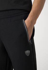 Svarta joggingbyxor med dragkedjefickor, elastisk midja och en texturerad logotyp på vänstra låret. Slät tyg med subtil lyster.