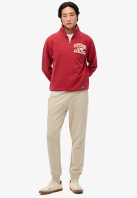Röd fleecepullover med en fjärdedels dragkedja och "SUPERDRY ATHLETICS" tryckt i krämfärg, tillsammans med beiga joggers och vita sneakers.