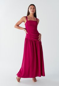 Vestido maxi fucsia con corpiño fruncido, tirantes anchos y falda recogida. Textura de tela lisa y brillo sutil. Sandalias planas con tiras.