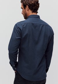 Donkerblauwe denim overhemd met een kraag, lange mouwen en knoopmanchetten. De achterkant heeft een gladde textuur met subtiele stikseldetails.