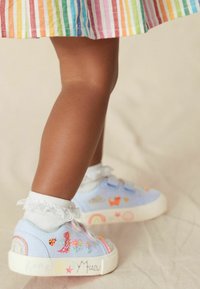 Světle modré plátěné tenisky s barevnými vyšívanými vzory včetně duhy, zvířat a hvězd; bílé volánkové kotníkové ponožky.